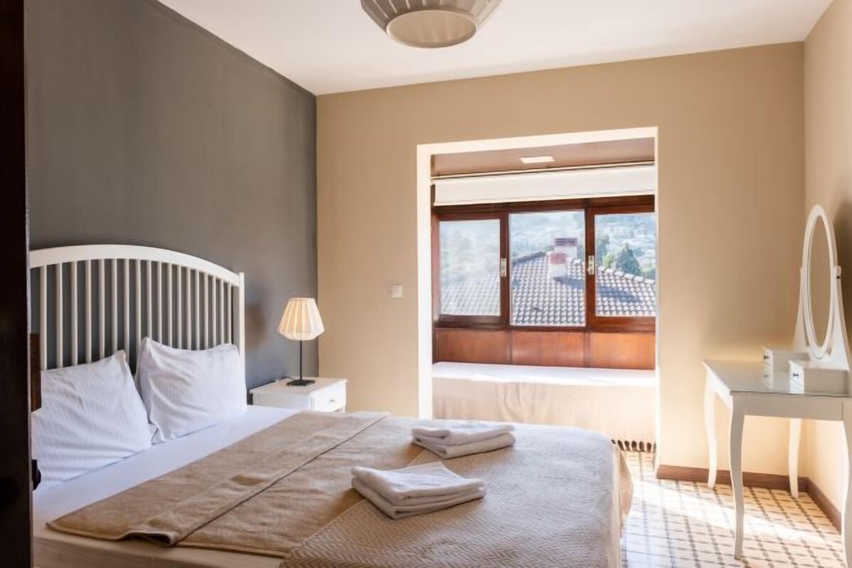 Un dormitorio con cama, mesita de noche con lámpara, ventana con vistas y espejo sobre un pedestal.
