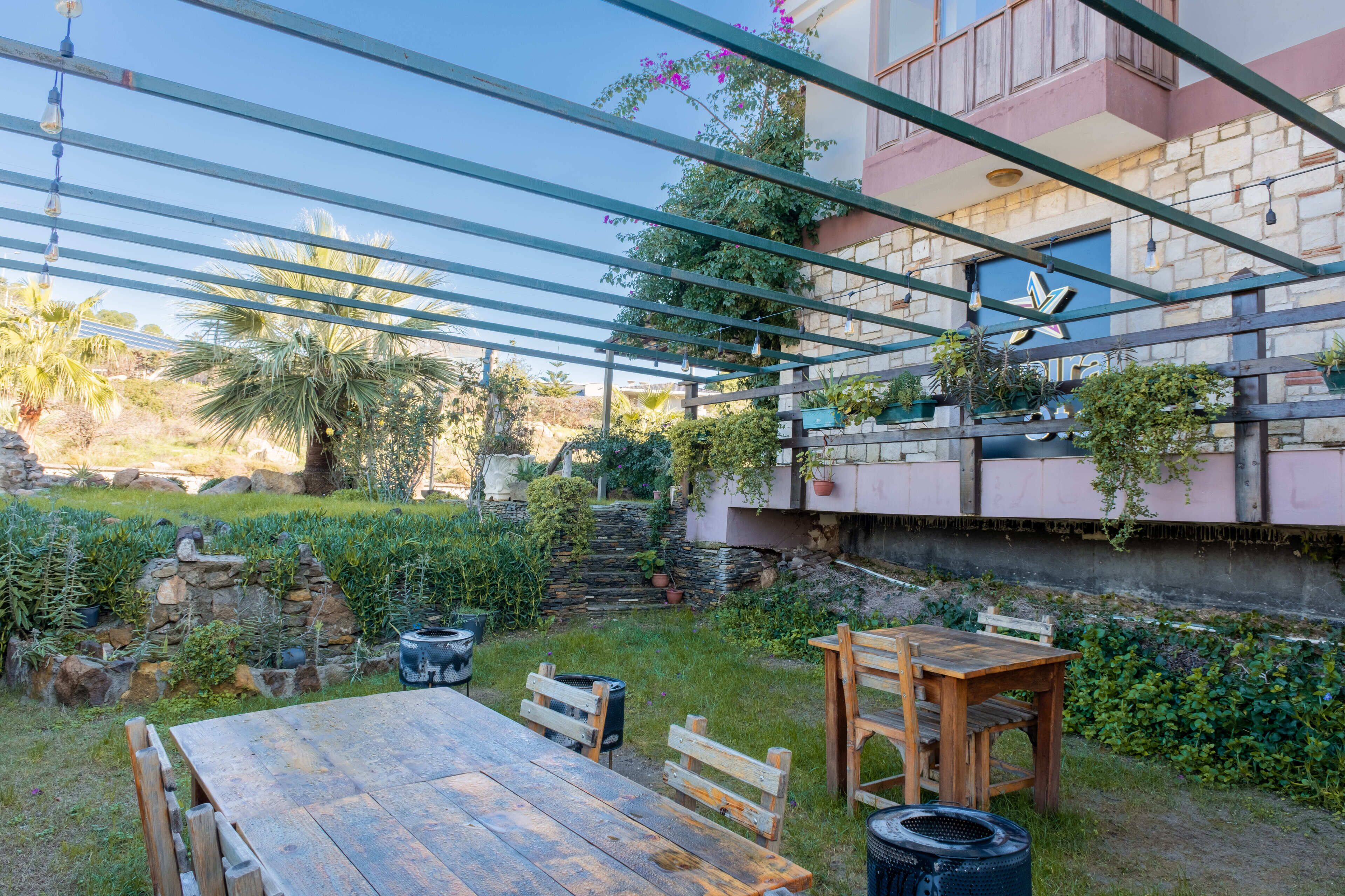 A patio area with a wooden table and benches, surrounded by greenery and a stone wall.