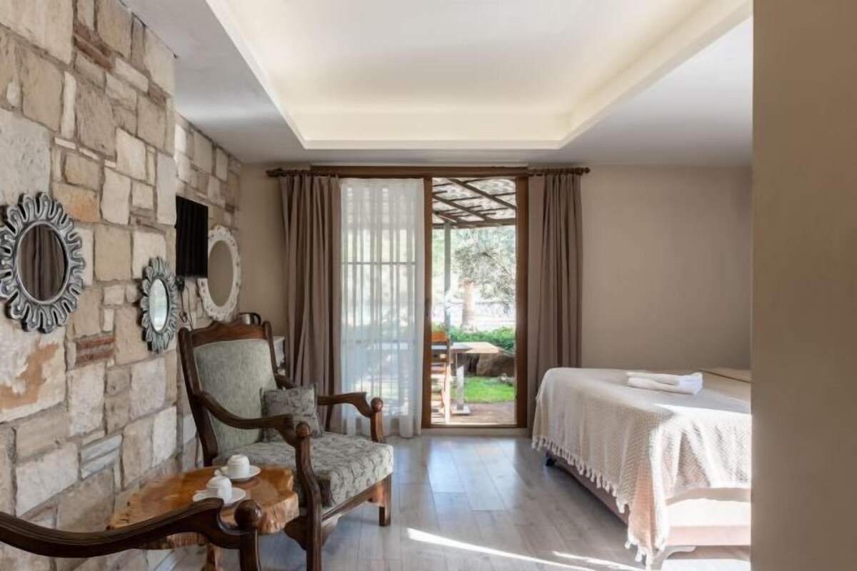 A room with a stone wall, a wooden chair, a bed with a white bedspread, and a table with a cup and saucer.