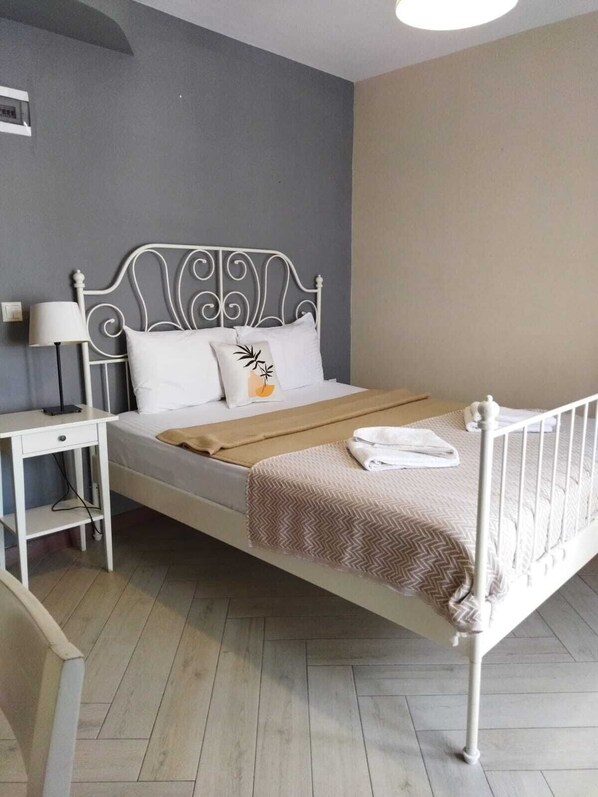 A single bed with a metal frame, white bedding, and a brown throw blanket. There is a bedside table with a lamp and a power outlet. The room has a grey wall and a wooden floor. - Espira by Antioch (Urla)