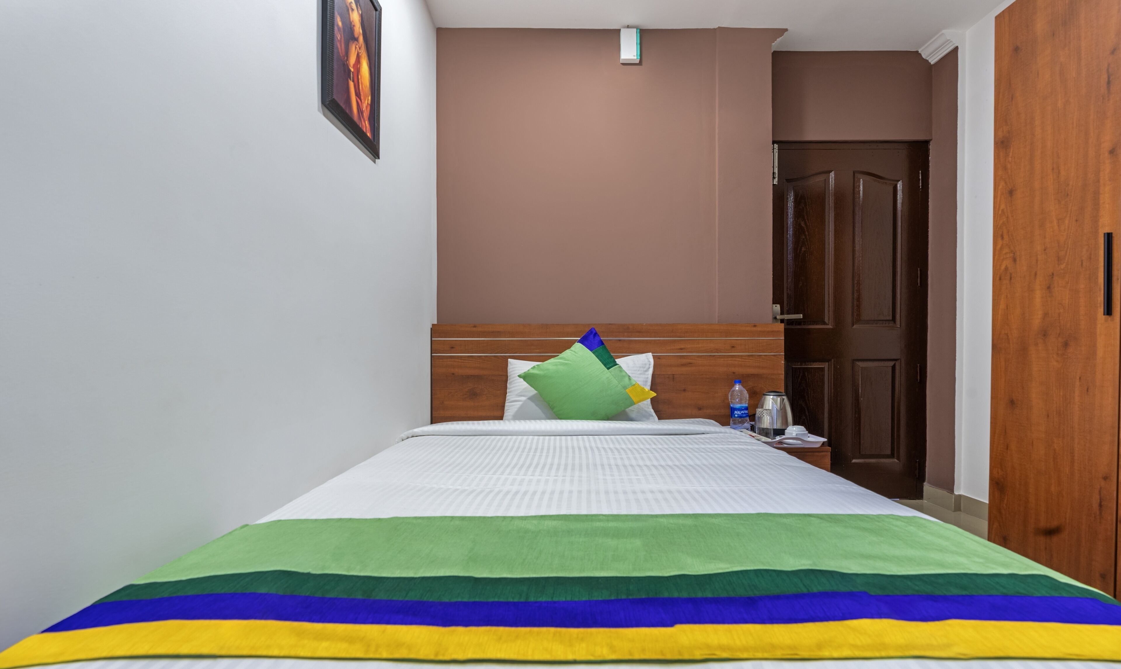 Una habitación de hotel con una cama, cabecera de madera, una puerta marrón y una pared blanca con un cuadro enmarcado.