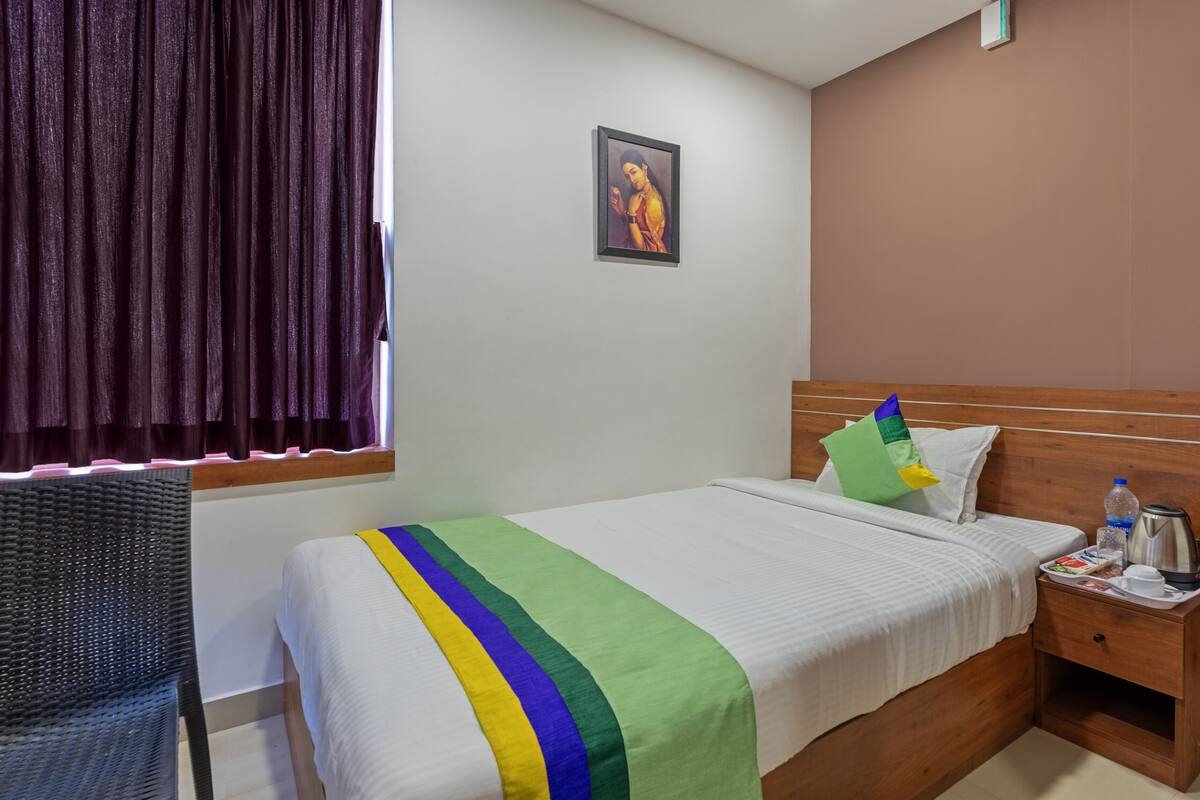 Quarto de hotel com cama, mesa de cabeceira, cadeira, janela com cortinas e um quadro na parede.