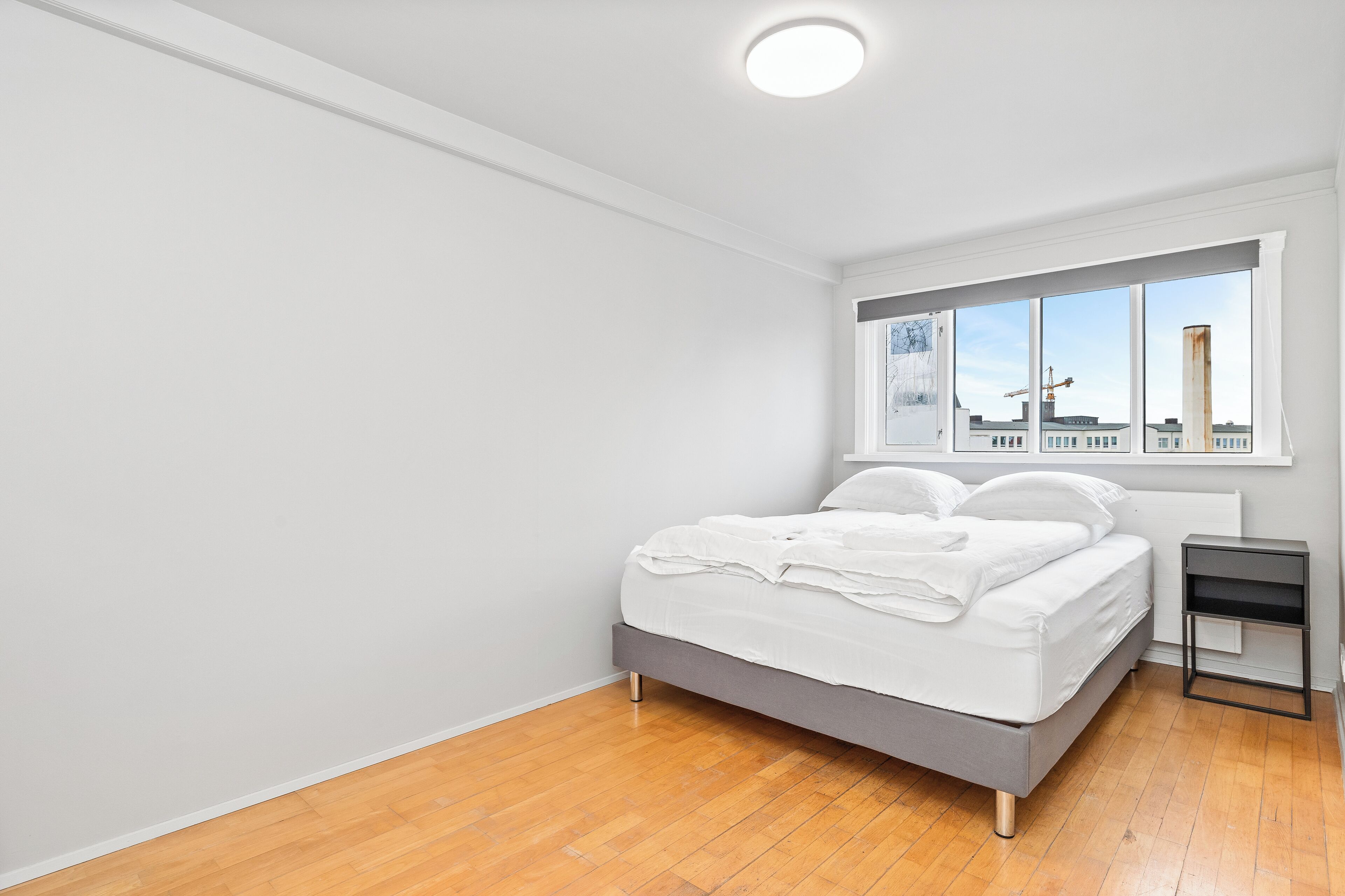 Un dormitorio con cama, mesita de noche, ventana con vista a edificios e iluminación en el techo.