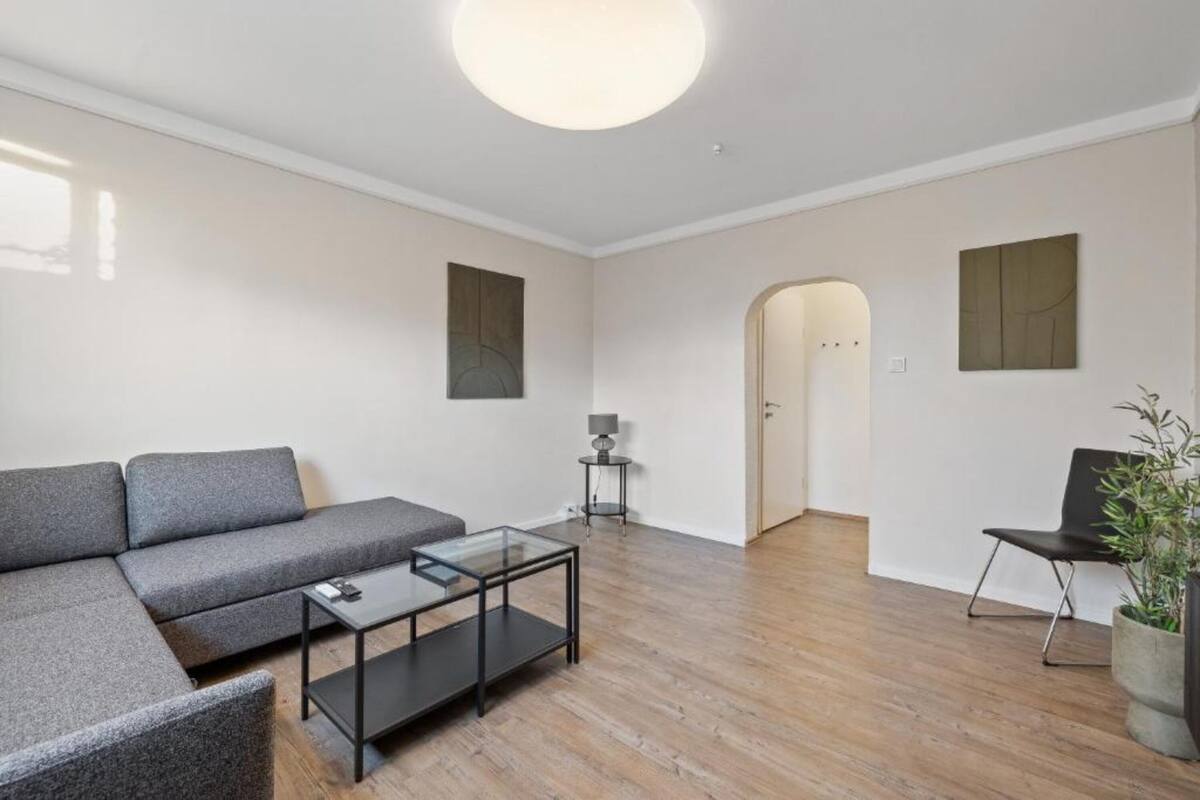 Een moderne woonkamer met een grijze hoekbank, een glazen salontafel en een zwarte fauteuil. Er staat een klein bijzettafeltje met een lamp en een potplant in de hoek.