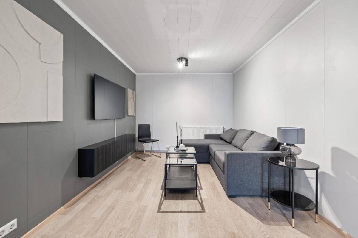 Een moderne woonkamer met een grijze bank, een salontafel, een bijzettafeltje en een flatscreen televisie aan de muur.