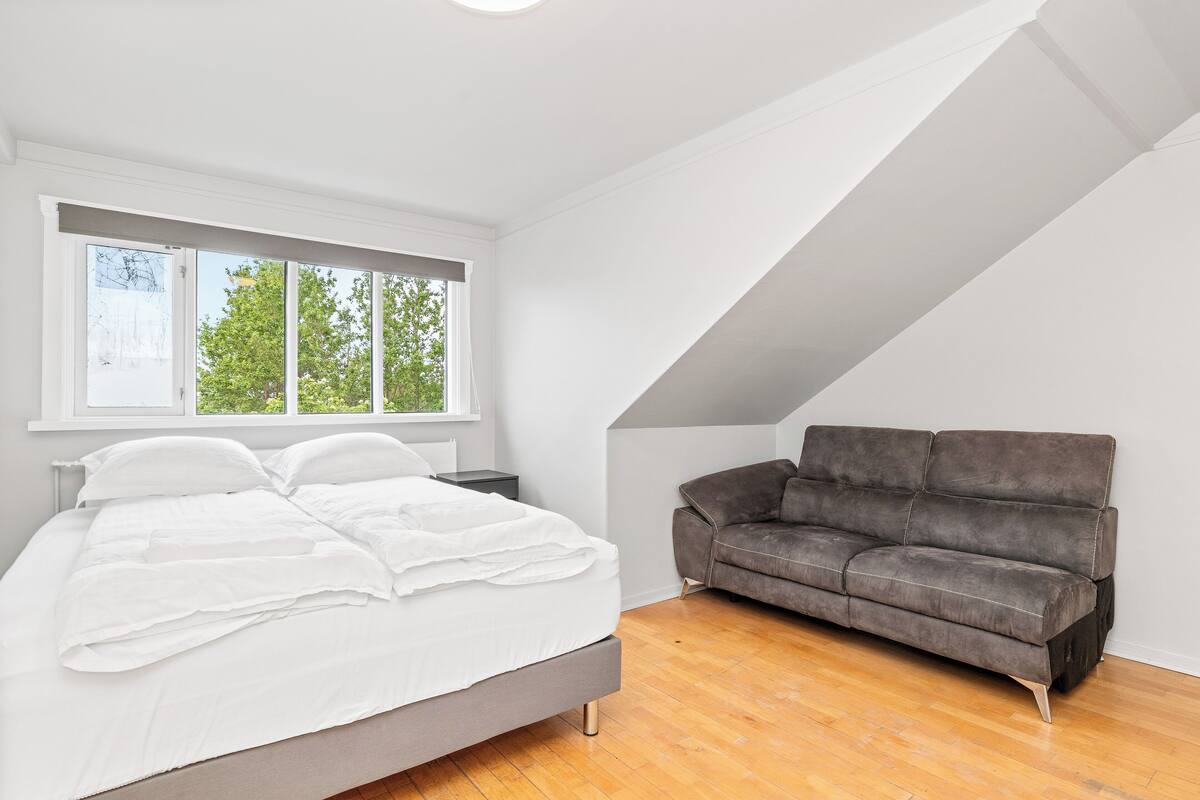 Een slaapkamer met een bed, een grijze bank, een raam met uitzicht op bomen en een schuine kap.