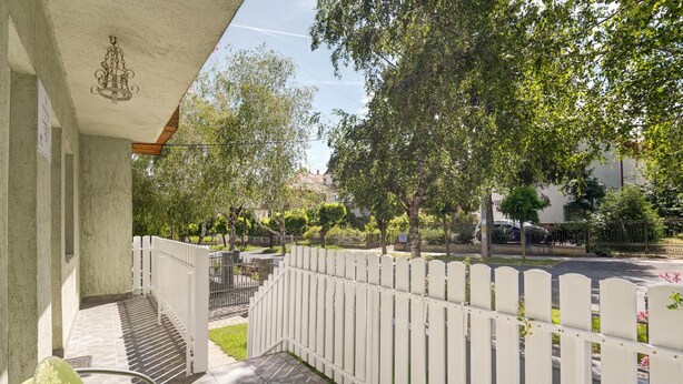 Ein Balkon mit weißem Zierzaun, einer hängenden Lampe und Blick auf eine von Bäumen gesäumte Straße.