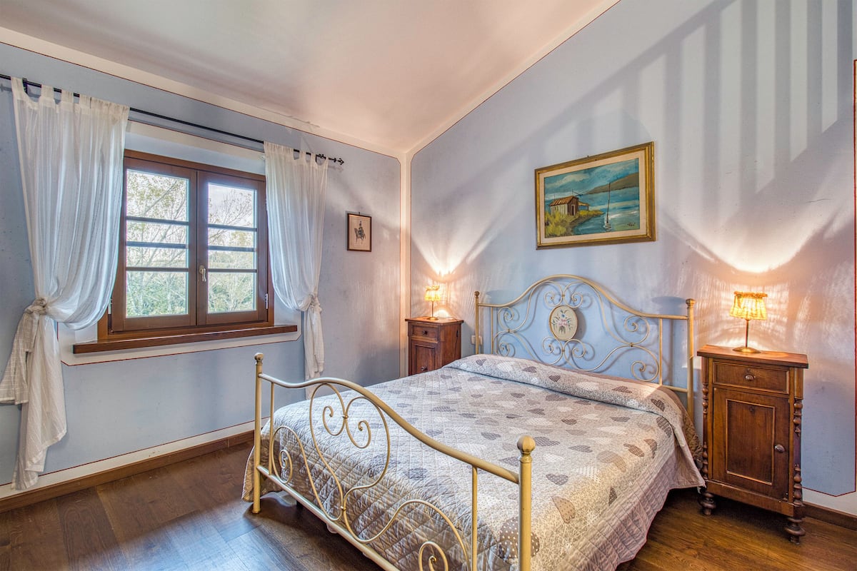 A bedroom with a bed, two bedside tables, a window with curtains, a painting on the wall, and a wooden floor.