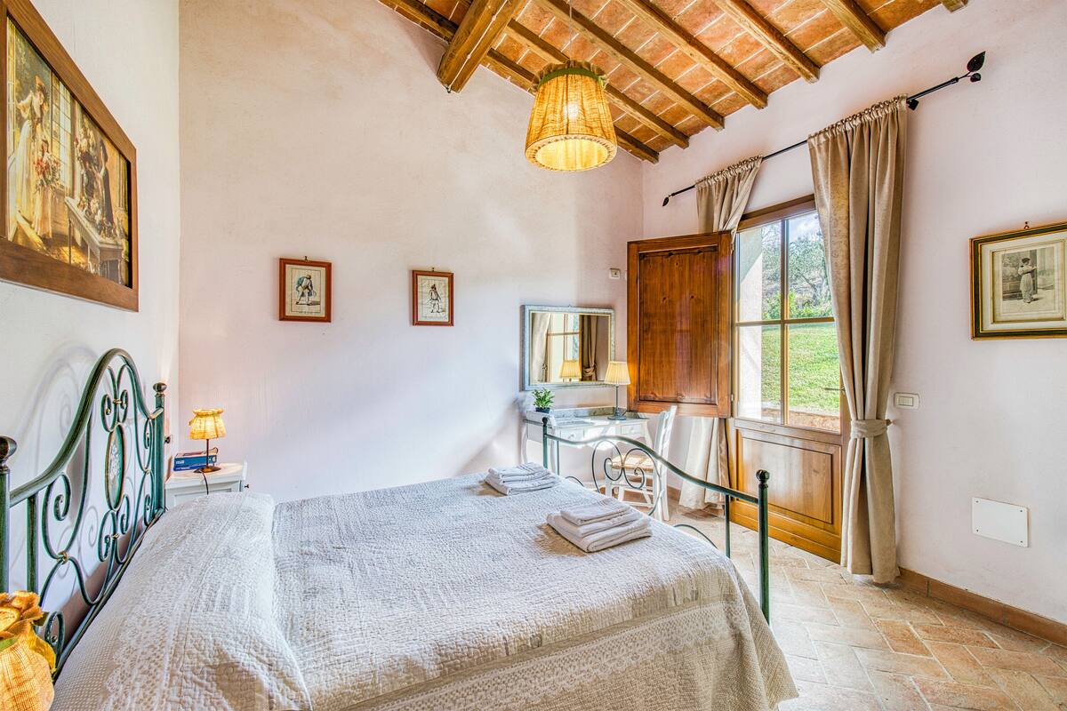 A bedroom with a bed, a desk, a window with curtains, and a painting on the wall.