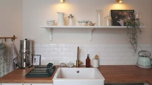 Une cuisine avec un évier blanc, un plan de travail en bois et un crédence carrelée blanche. On y trouve deux luminaires fixés au mur, une étagère avec divers ustensiles de cuisine et une bouilloire posée sur le plan de travail.