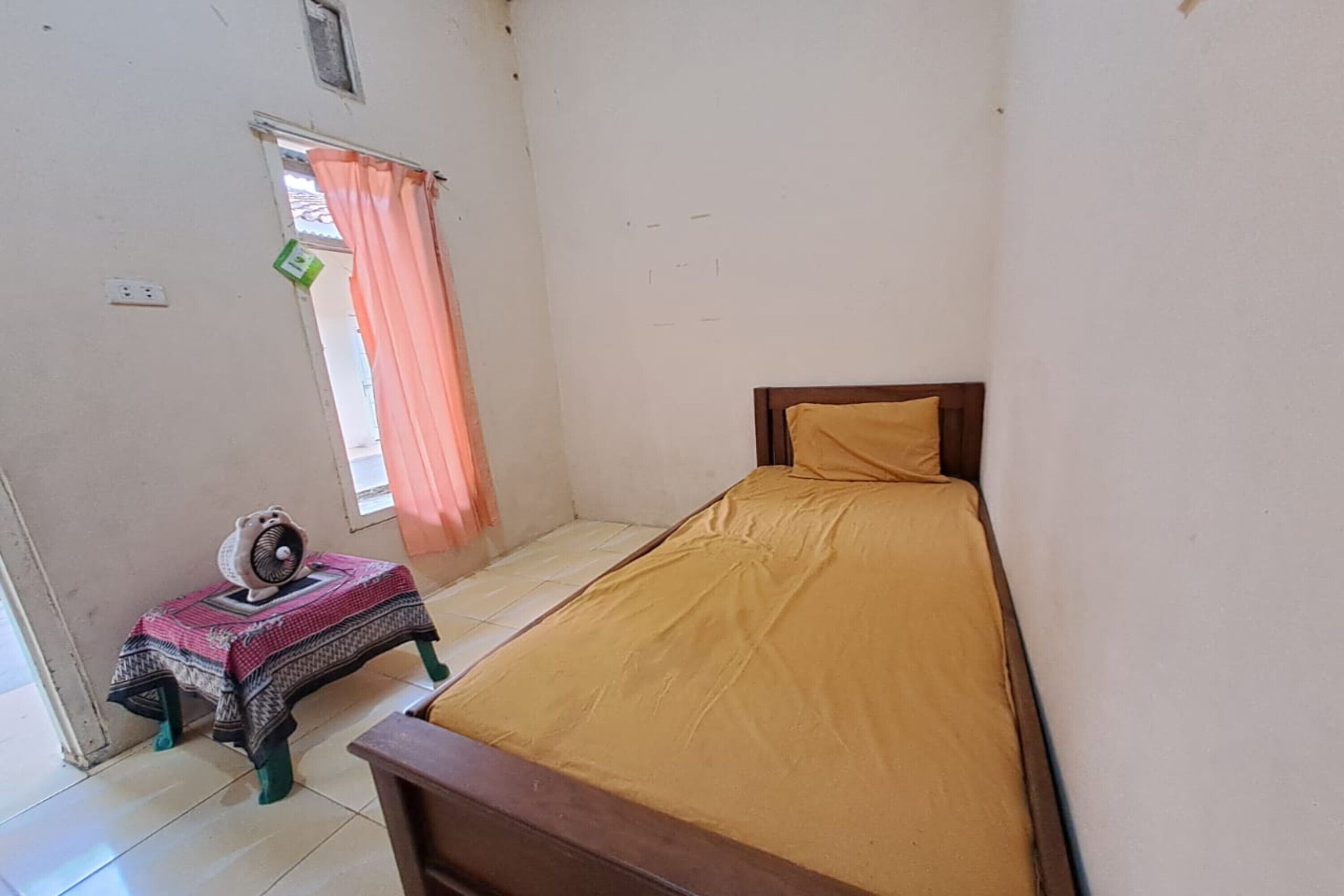 A small, single-bed room with a wooden headboard, a small table with a fan, and a window with a curtain.