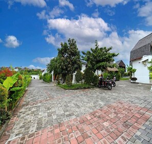 A paved driveway leading to a building with a thatched roof, surrounded by greenery and a clear blue sky. - Belvilla Seaside Hermitage Bungalow Canggu Beach (Nusa Dua)