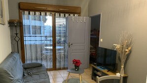 A living room with a grey sofa, a small table with a vase of flowers, a television, and a door leading to a balcony with a table and chairs. - Apparemment des Cèdres (Uzès)