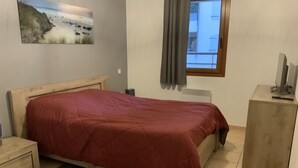 A bedroom with a bed, a nightstand, a bedside table, a radiator, a window with curtains, and a painting on the wall. - Apparemment des Cèdres (Uzès)