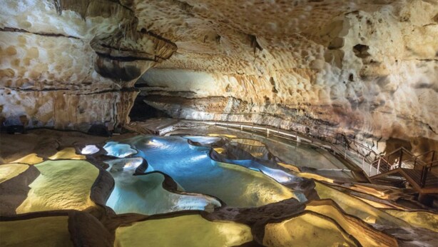 A cave with stalactites and stalagmites, a water pool, and a metal railing. - Apparemment des Cèdres (Uzès)
