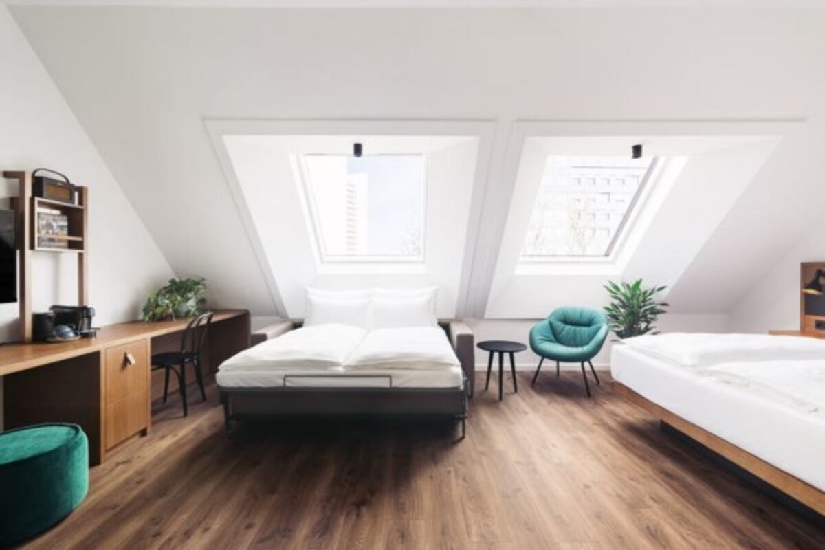 Un dormitorio moderno con cama elevada, un escritorio y una silla. Hay tragaluces sobre la cama y una pequeña mesa con una silla.