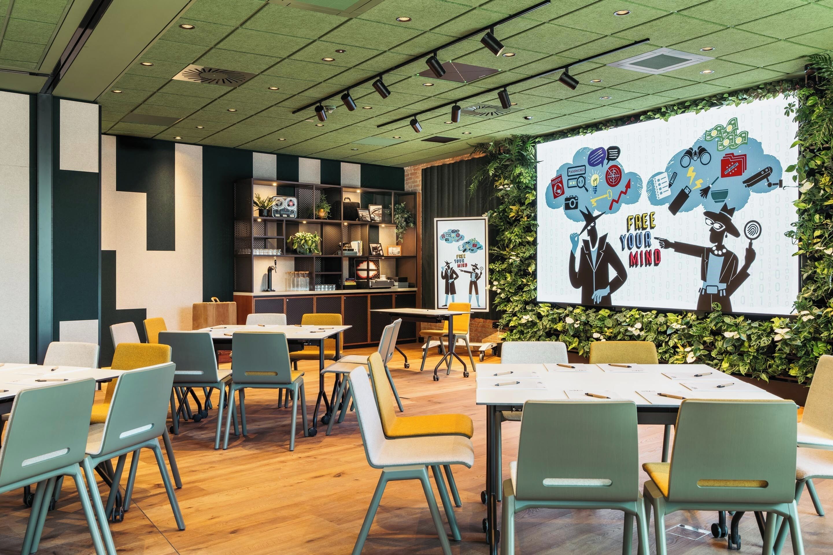 Una sala de conferencias moderna con mesas y sillas, una pantalla de proyector y una pared verde con un mural.