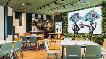 Una sala de conferencias moderna con mesas y sillas, una pantalla de proyector y una pared verde con un mural.