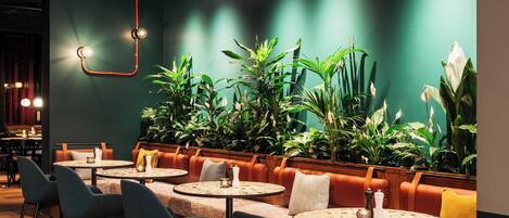 A modern restaurant with round tables, cushioned booths, and a variety of green plants.