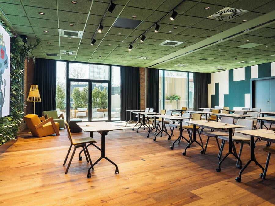 Una sala de conferencias moderna con suelo de madera, mesas y sillas, ventanas amplias y una pantalla de proyector.