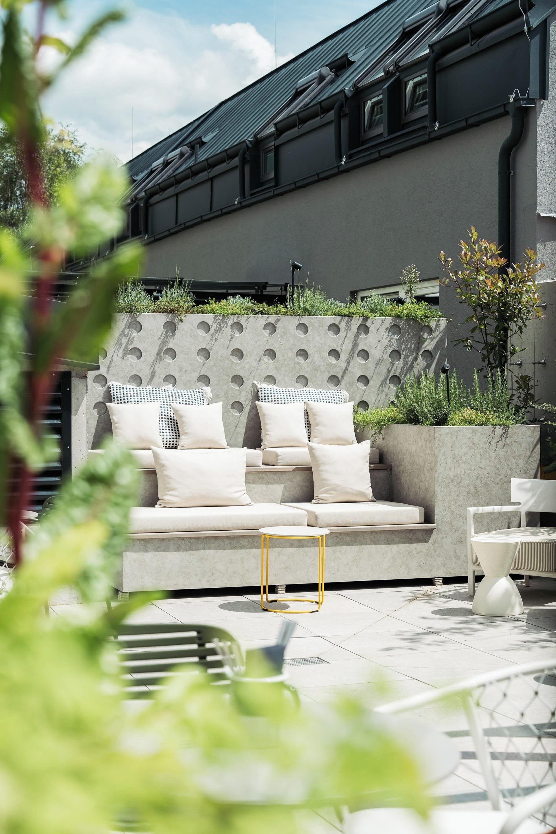 Un área de estar moderna al aire libre con sofá y sillones, un macetero de concreto con follaje, y un edificio con techo y ventanas oscuras al fondo.