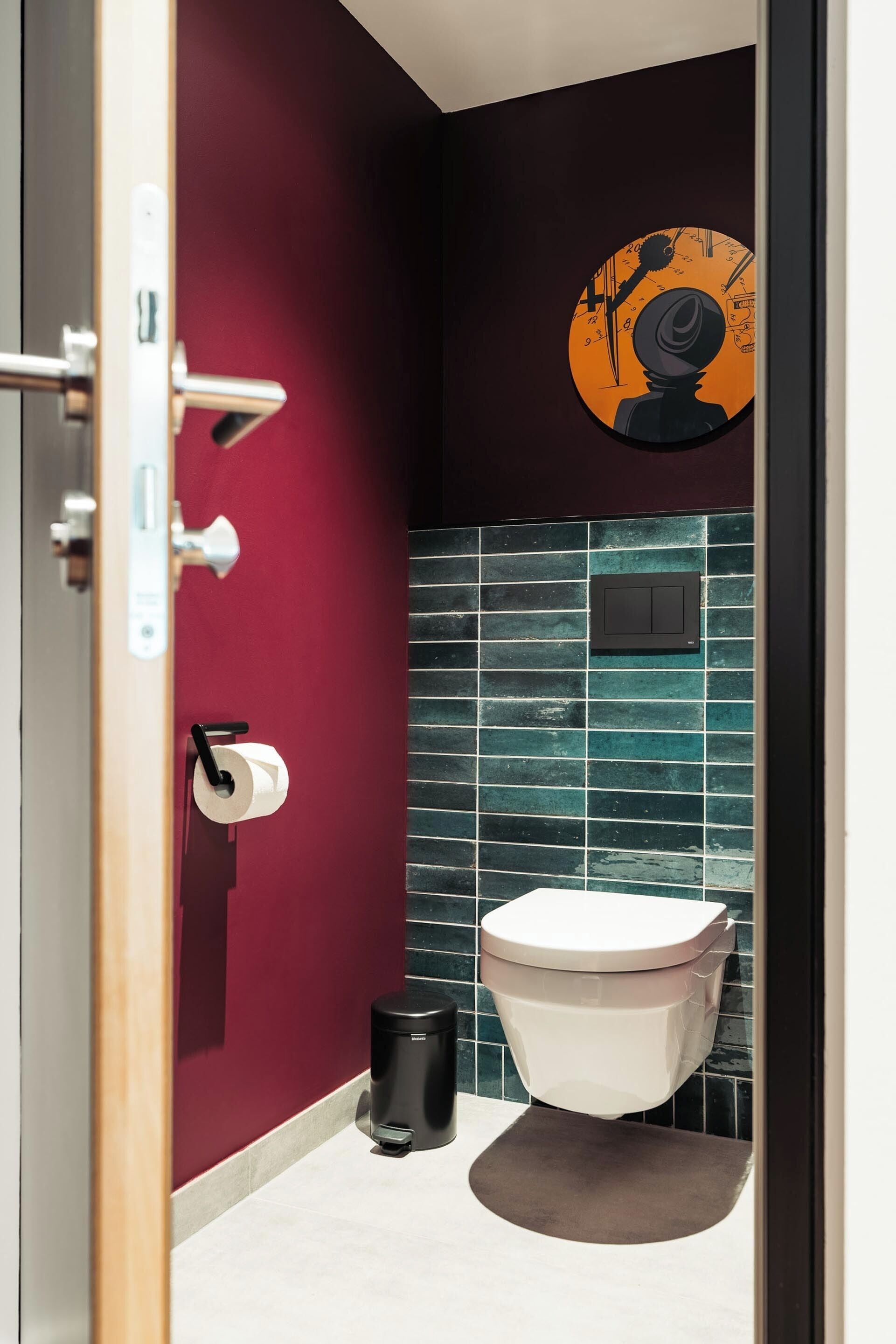 A bathroom with a toilet, a roll of toilet paper, a trash bin, and a wall-mounted light switch.