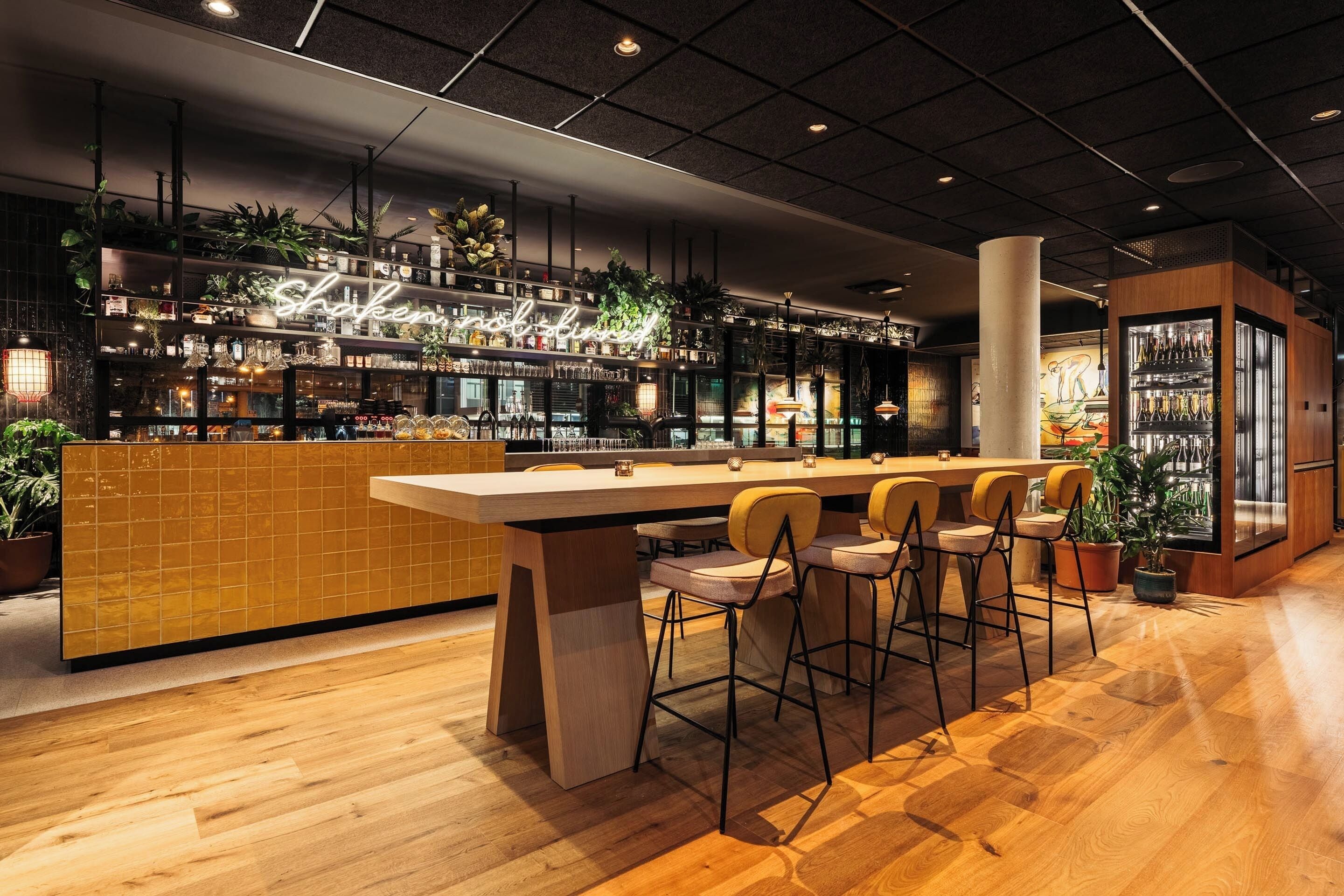 A modern bar area with a long counter, bar stools, and shelves stocked with bottles and plants.