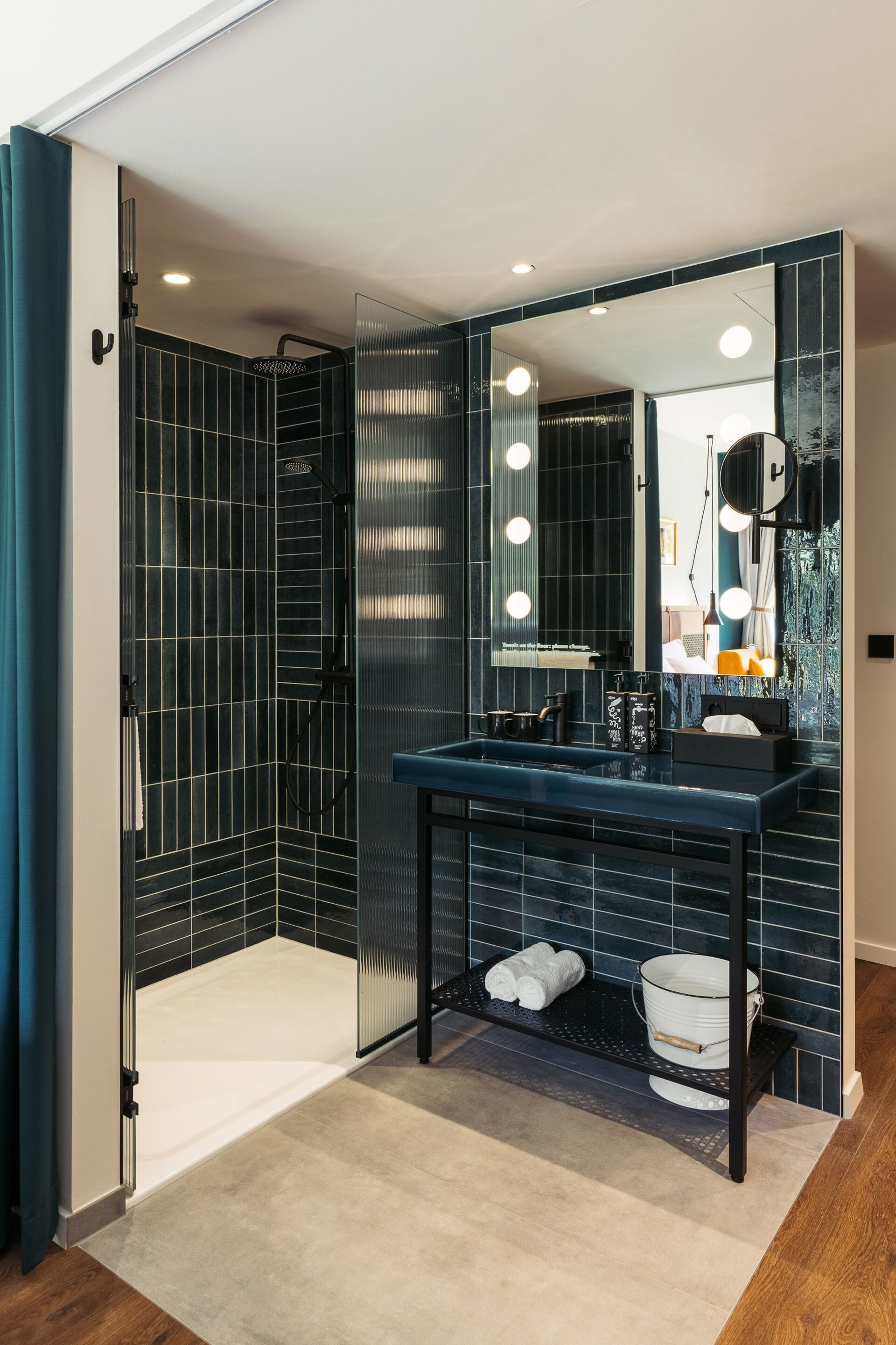 A modern bathroom with a dark tiled shower, a large mirror with lights, and a black countertop with a sink and towel rack.