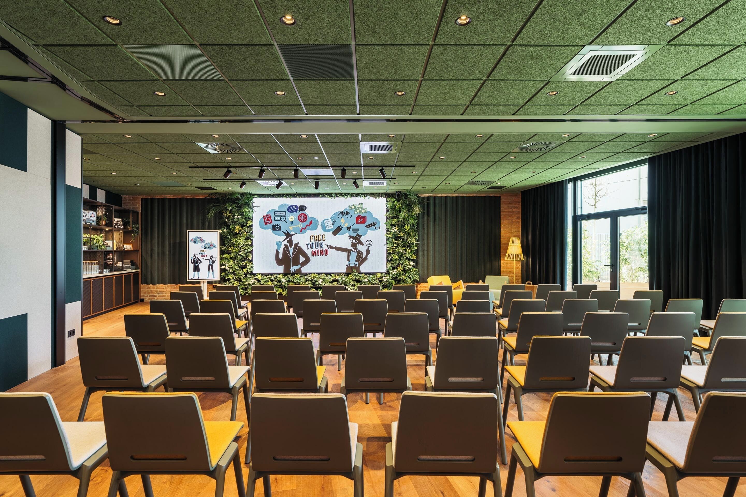 A conference room with rows of chairs and tables, a large screen displaying a presentation, and a green ceiling with recessed lighting.
