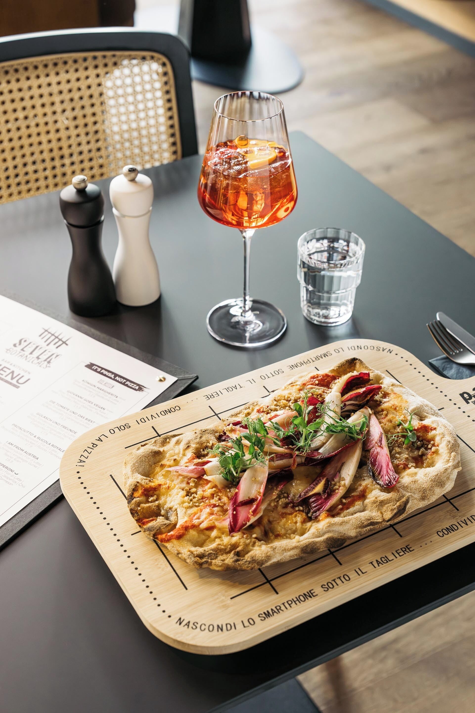 Ein Holzschnittbrett mit einer Pizza, ein Glas Rotwein und eine Speisekarte auf einem Tisch.