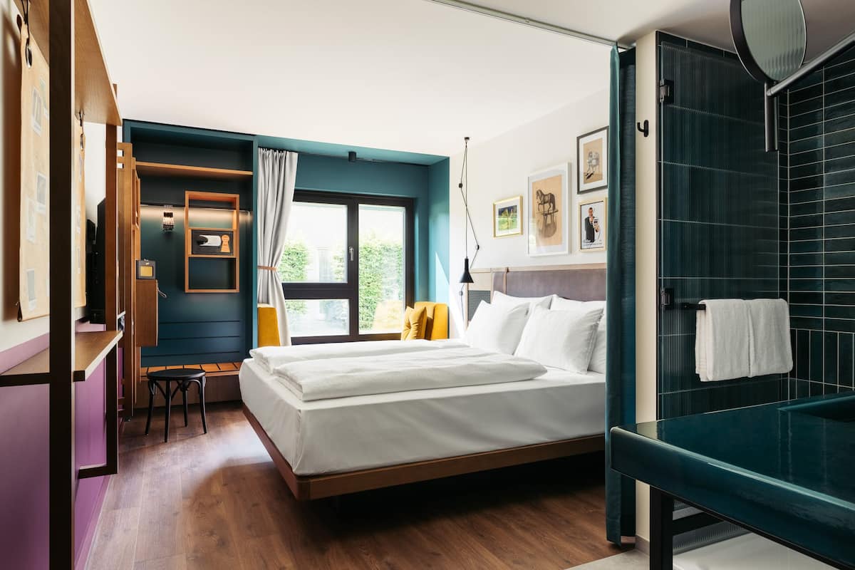Ein Hotelzimmer mit Bett, Schreibtisch, Stuhl, einem Fenster mit Vorhängen, einem Badezimmer mit Eckbadewanne und Bildern an den Wänden.