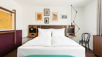 Een net opgemaakt bed met witte lakens, een groene plaids en een houten hoofdbord. Daarboven hangen ingelijste kunstwerken aan de muur en rechts staat een bureaulamp.