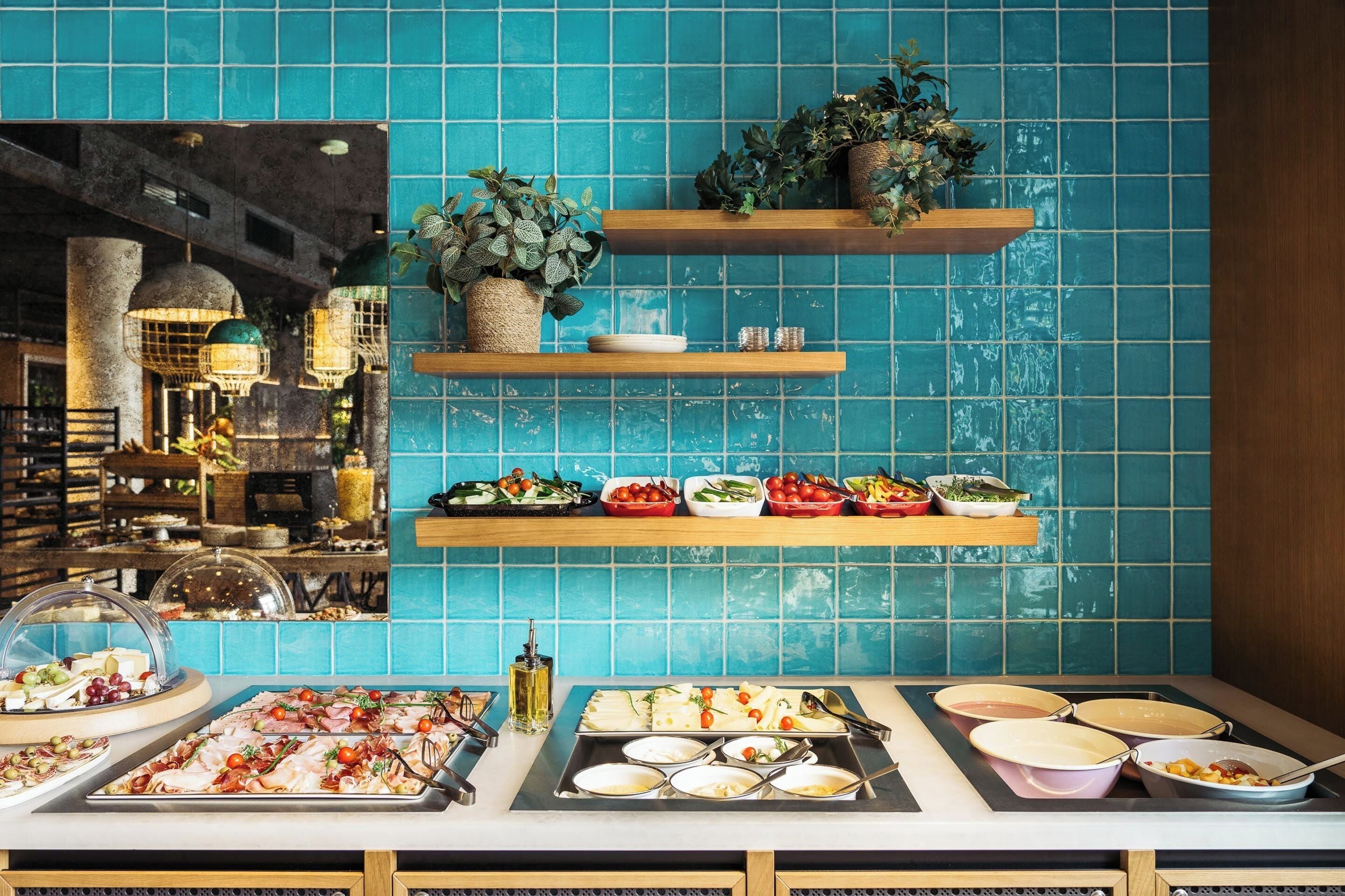 A buffet with various dishes and a blue tiled wall with wooden shelves holding plants.
