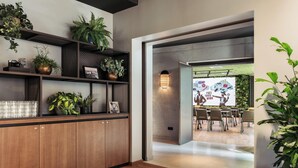 Een modern interieur met houten vloeren, een houten kast en planken met planten. Er hangt een lamp aan de muur en een deur leidt naar een eetgedeelte.