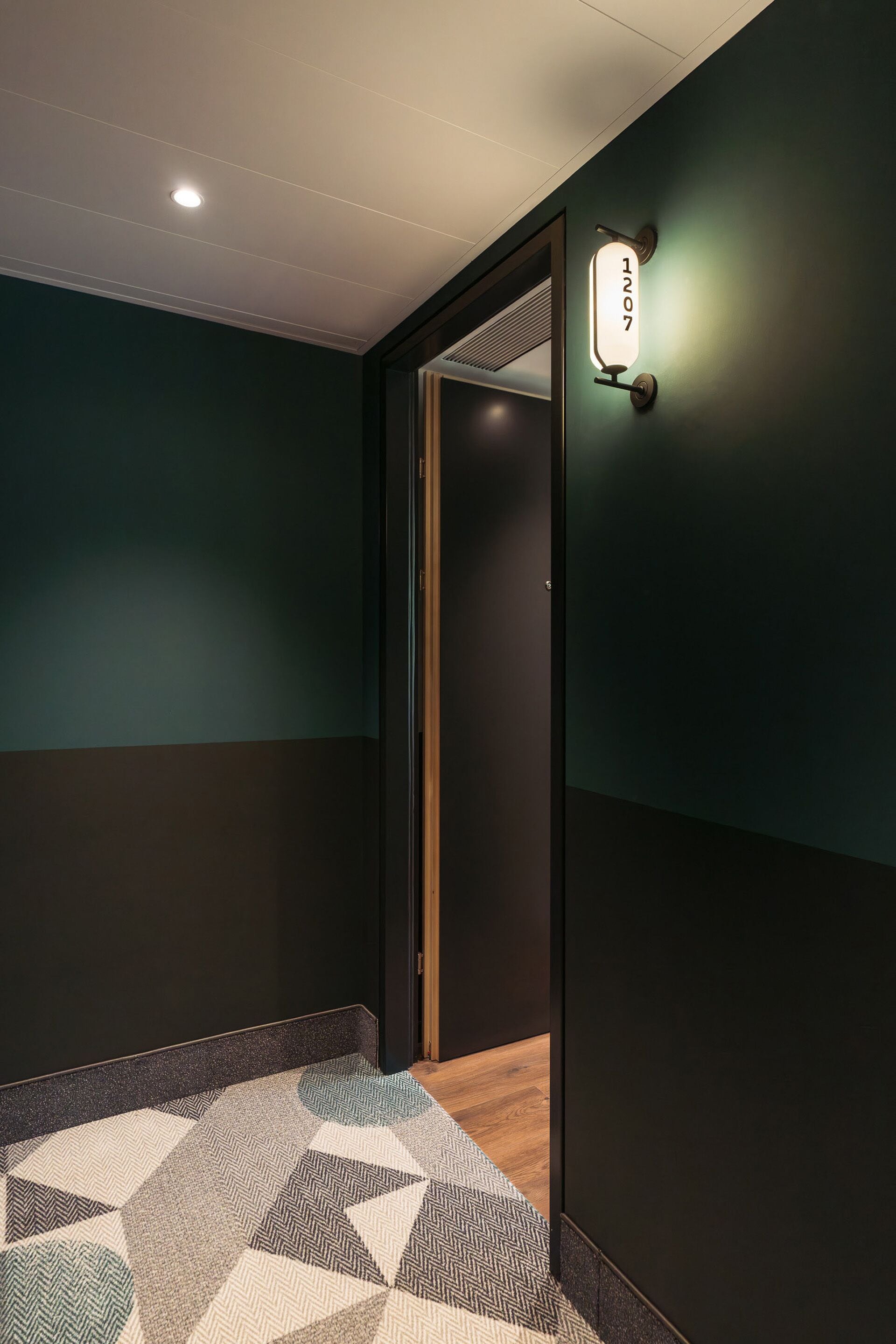 Un couloir avec un tapis à motifs géométriques, un luminaire fixé au mur et une porte portant un numéro.