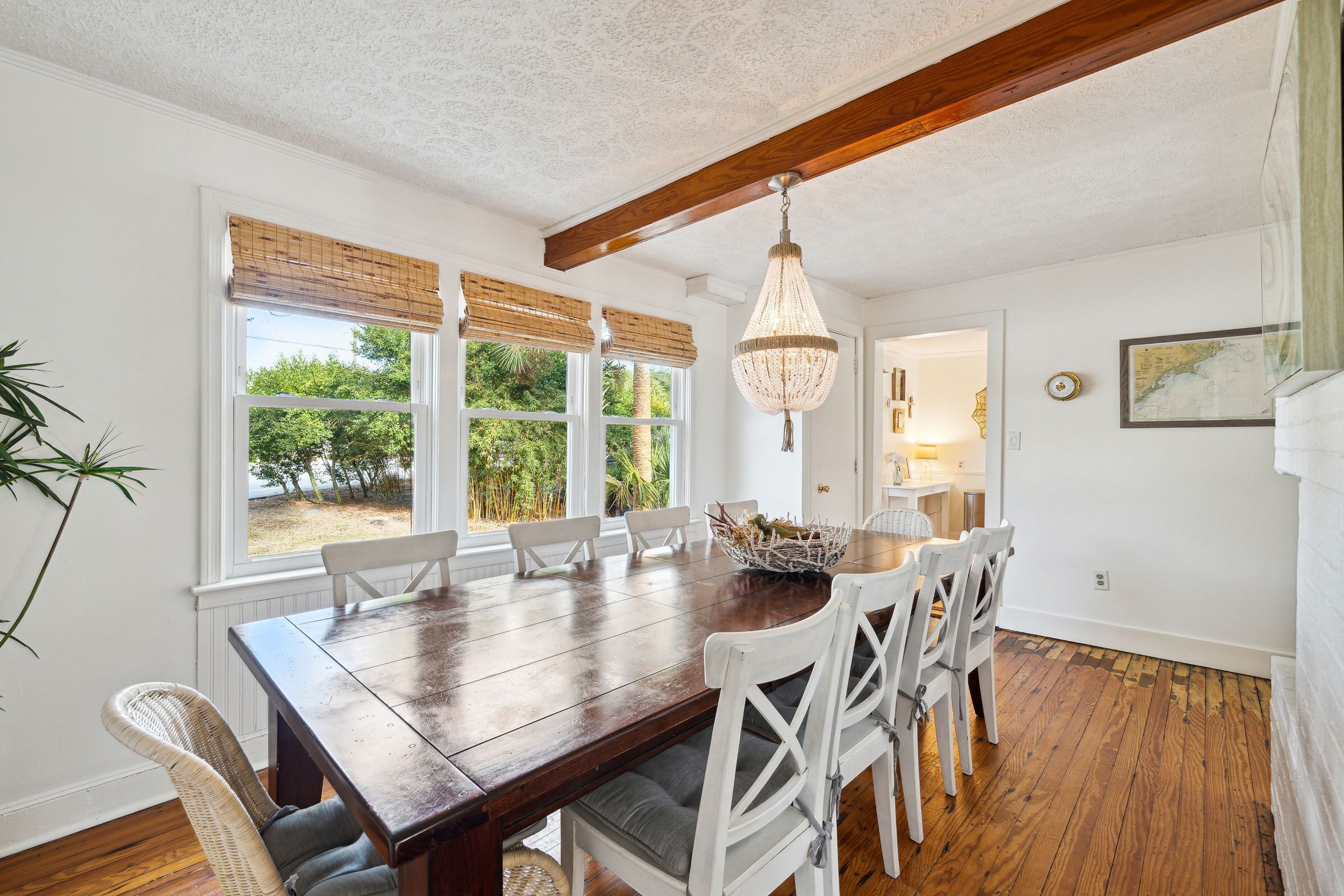 Une salle à manger avec une table en bois, des chaises blanches, un lustre suspendu et de grandes fenêtres équipées de stores.