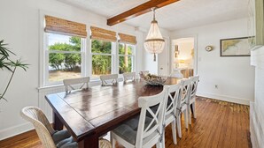 Comedor con mesa de madera, sillas blancas, lámpara de araña colgante y ventanas grandes con persianas.