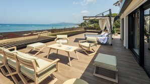 A sunlit outdoor seating area with lounge chairs and a table, overlooking a beach with boats and hills in the distance. - Cala Beach Resort (Castiglione della Pescaia)