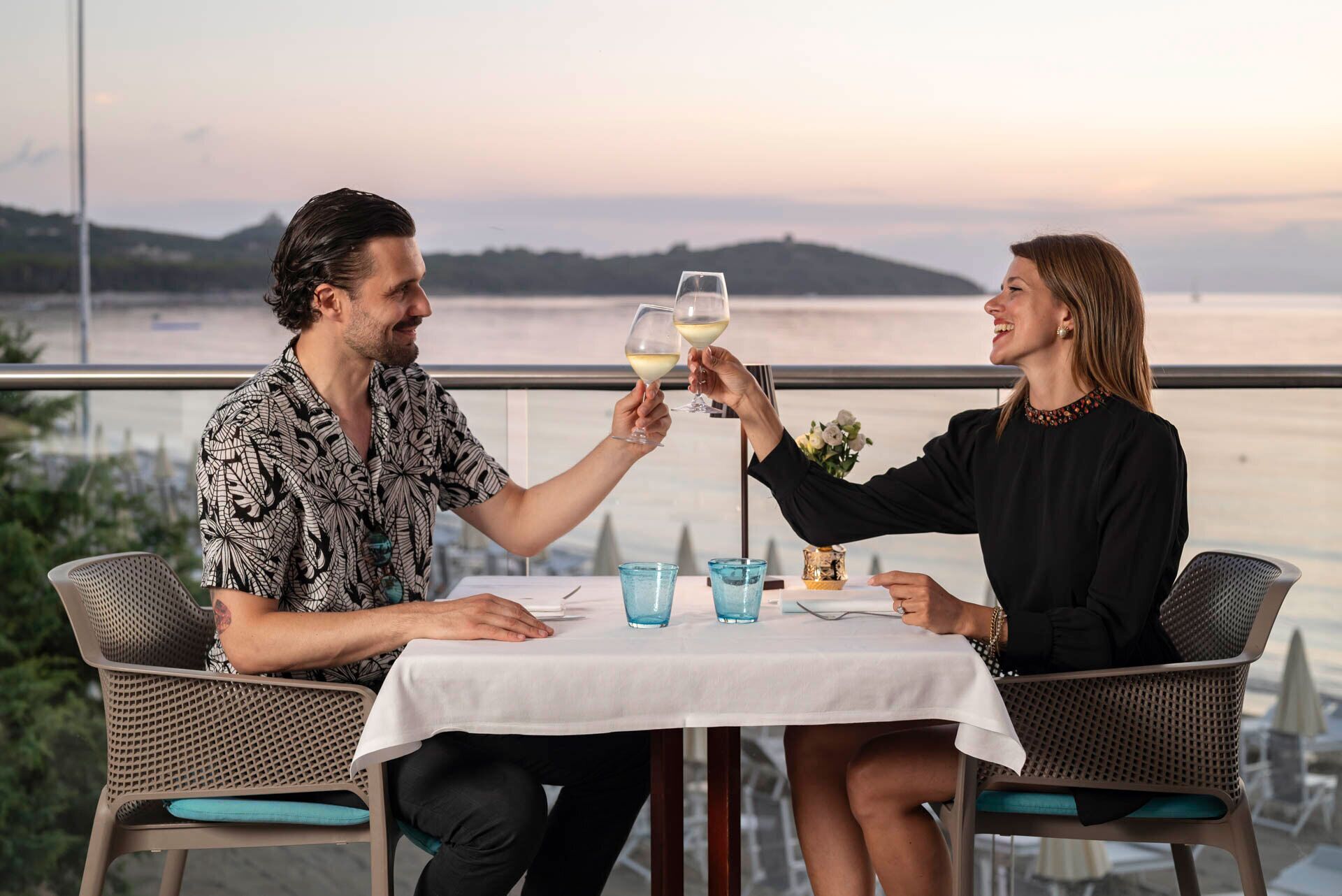 Deux personnes trinquent avec des verres de vin, assises à une table d’où l’on peut admirer la mer et les collines.