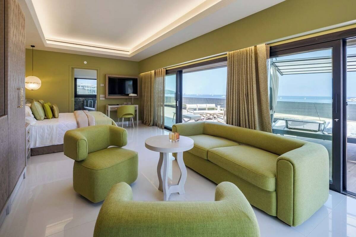 Habitación de hotel moderna con una cama grande, un sofá, dos sillones, una mesita y un televisor. La habitación tiene ventanas amplias con vistas al mar.