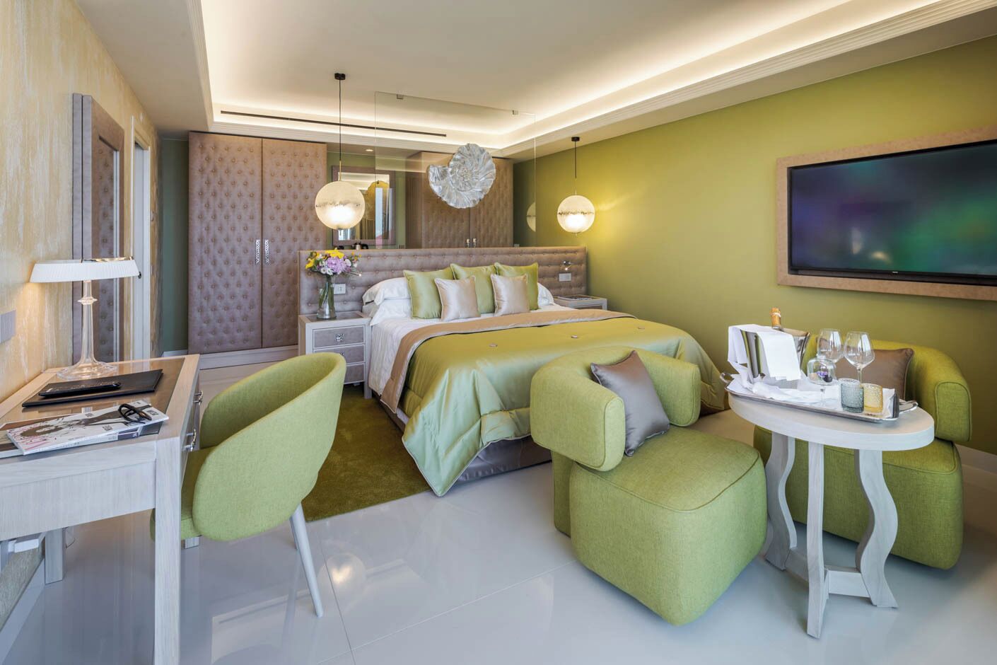 Ein modernes Hotelzimmer mit einem großen Bett, einem Schreibtisch mit Lampe, einem kleinen Tisch mit Tablett und grünen Polsterstühlen.