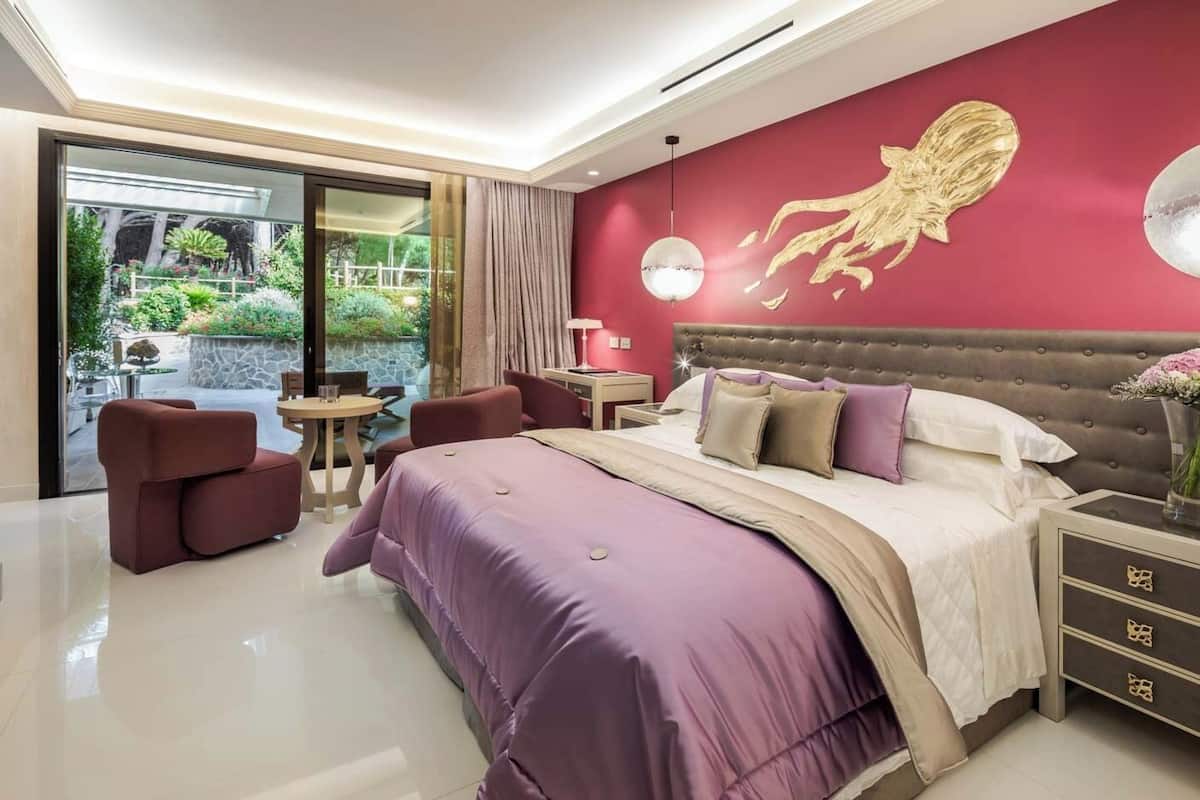 Un dormitorio moderno con una cama grande, una pared roja con diseño dorado, un vidrio que da a un área exterior y una pequeña mesa con sillas.