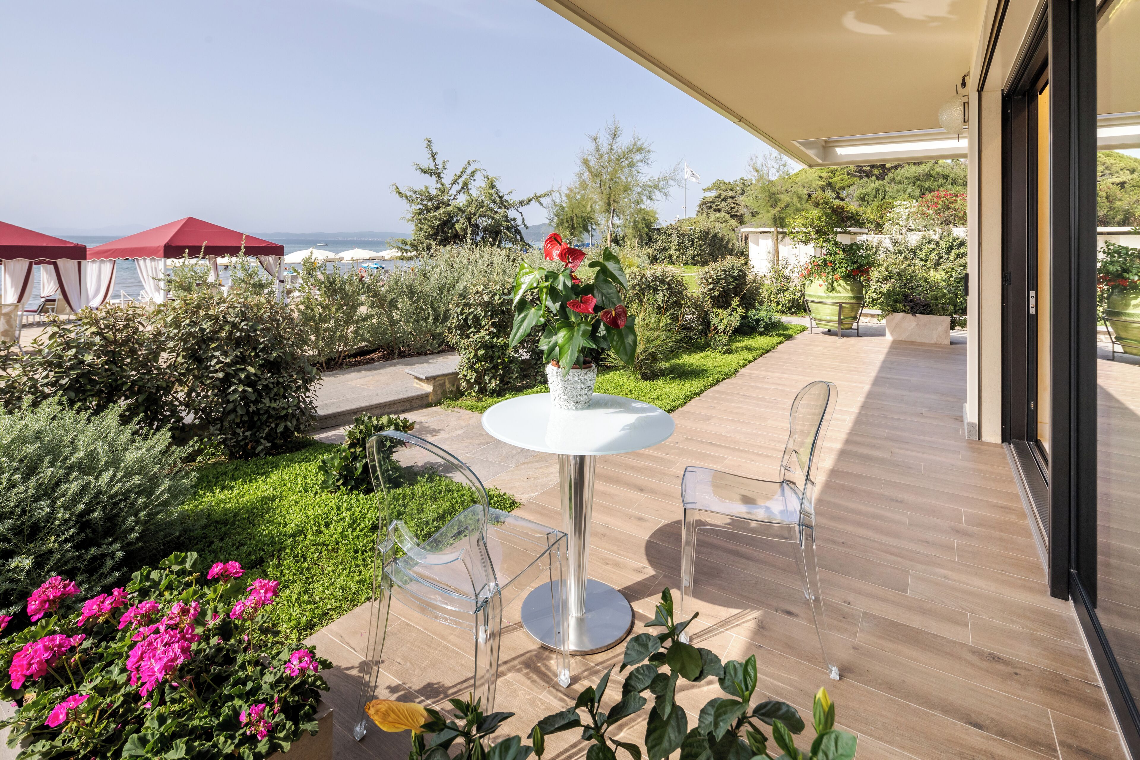 A patio with a round table and two chairs, surrounded by greenery and a view of the sea.