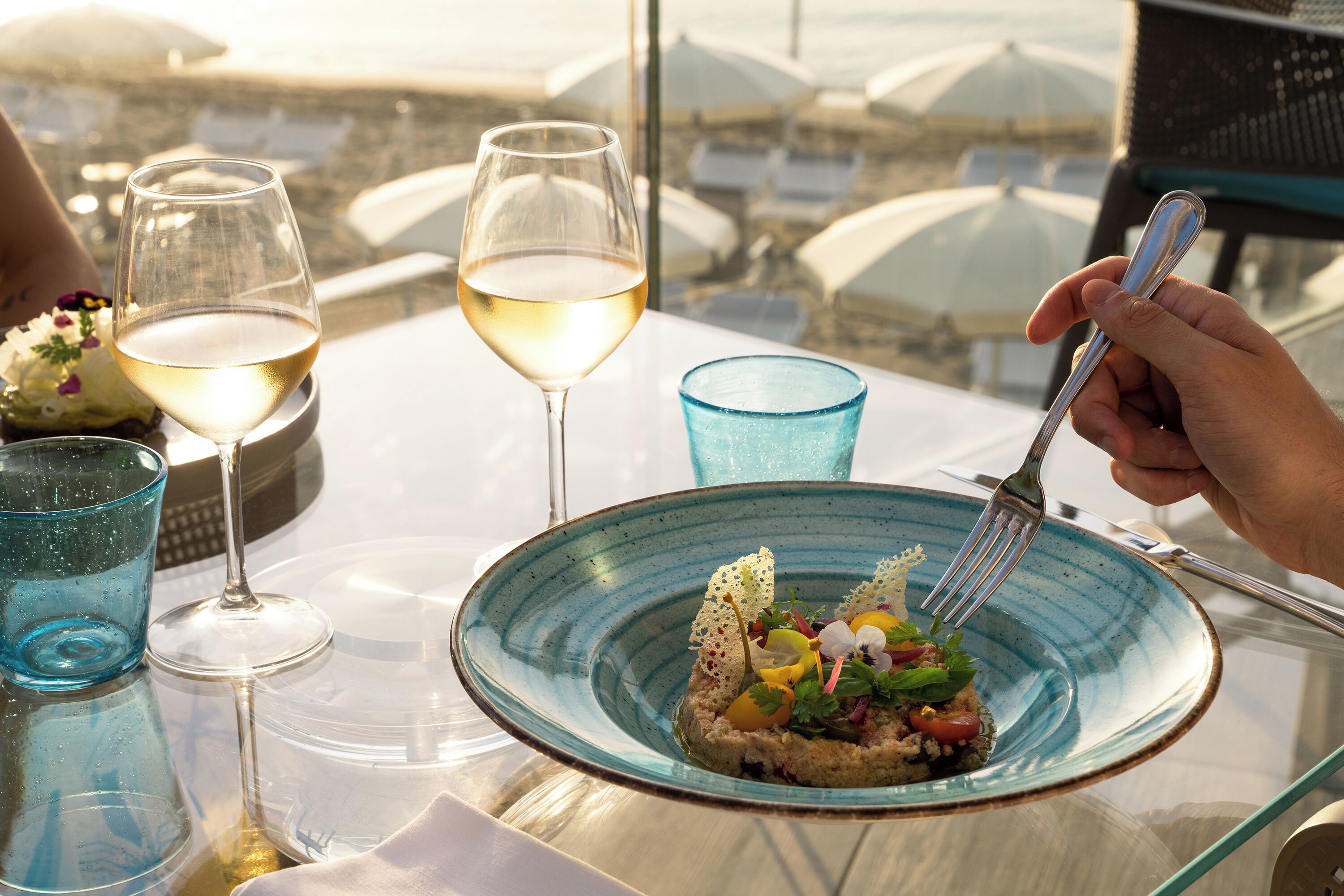 A table with a glass of white wine, a blue glass, and a plate of food.