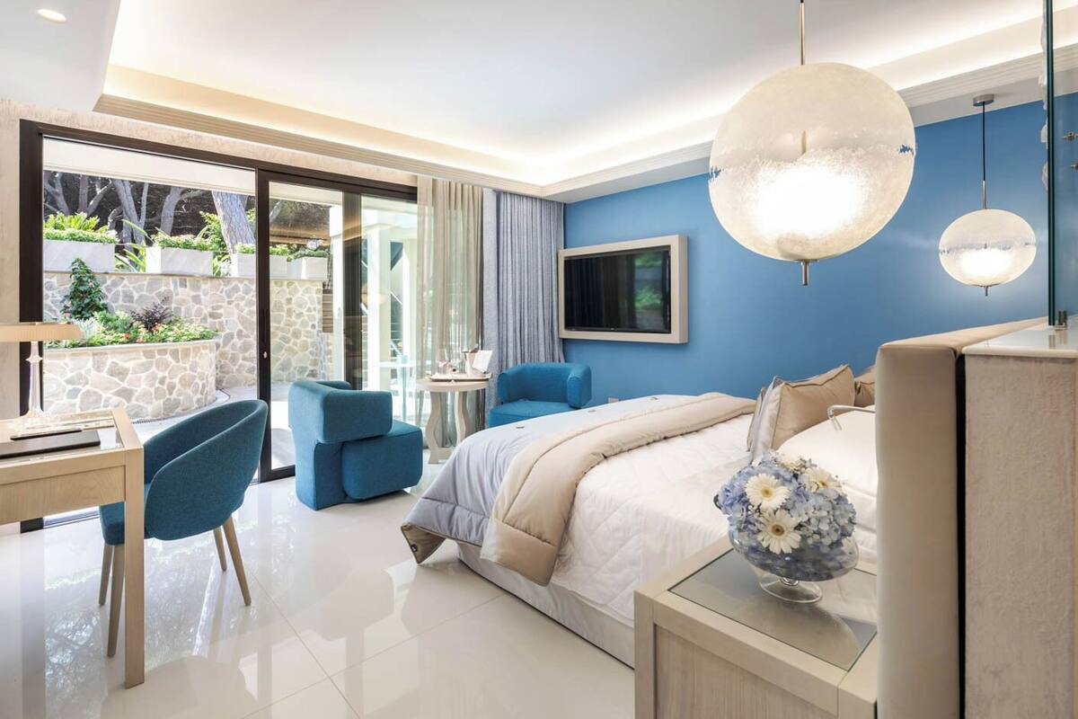 Habitación de hotel moderna con una cama grande, un escritorio y una silla. Hay un televisor montado en la pared y un ventanal con vistas a zonas verdes.