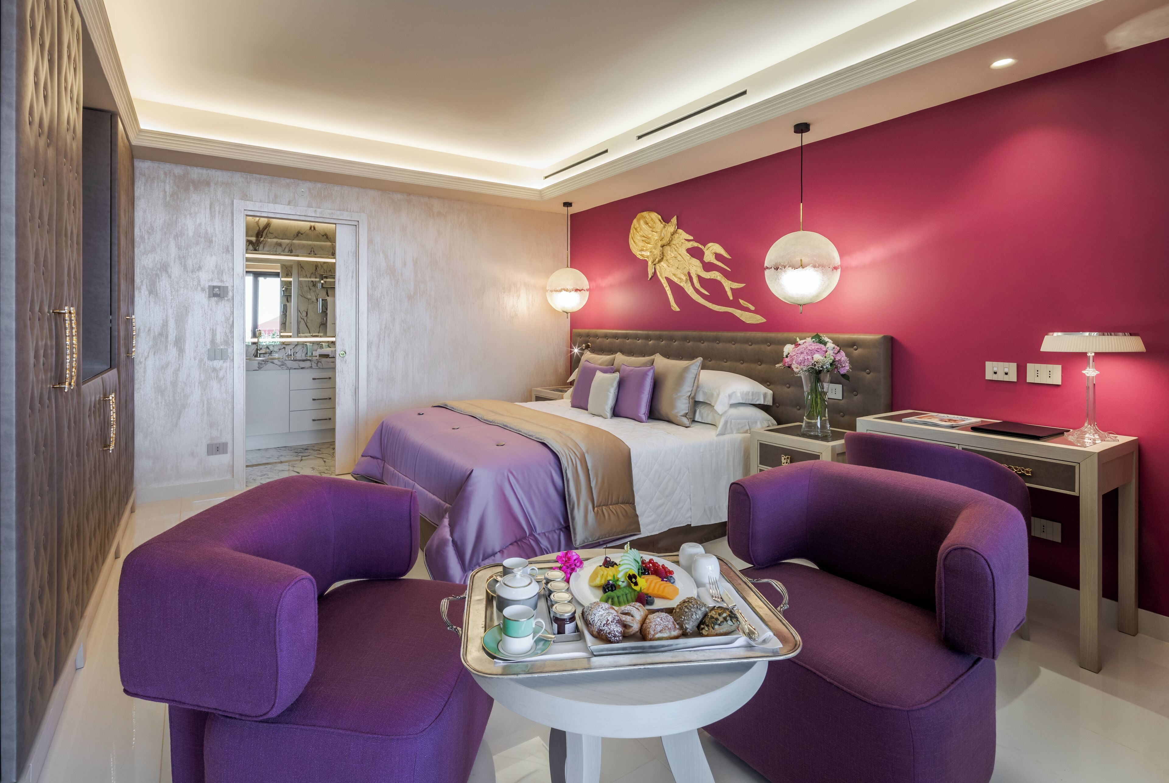 A modern hotel room with a large bed, two purple armchairs, a small table with breakfast, and a vibrant red accent wall.