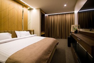 Hotelkamer met een groot bed, een houten hoofdbord, een flatscreen televisie, een bureau en een groot raam met gordijnen.