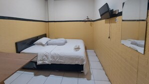 A small, compact room with a bed, a wooden desk, a mirror, and a TV mounted on the wall.