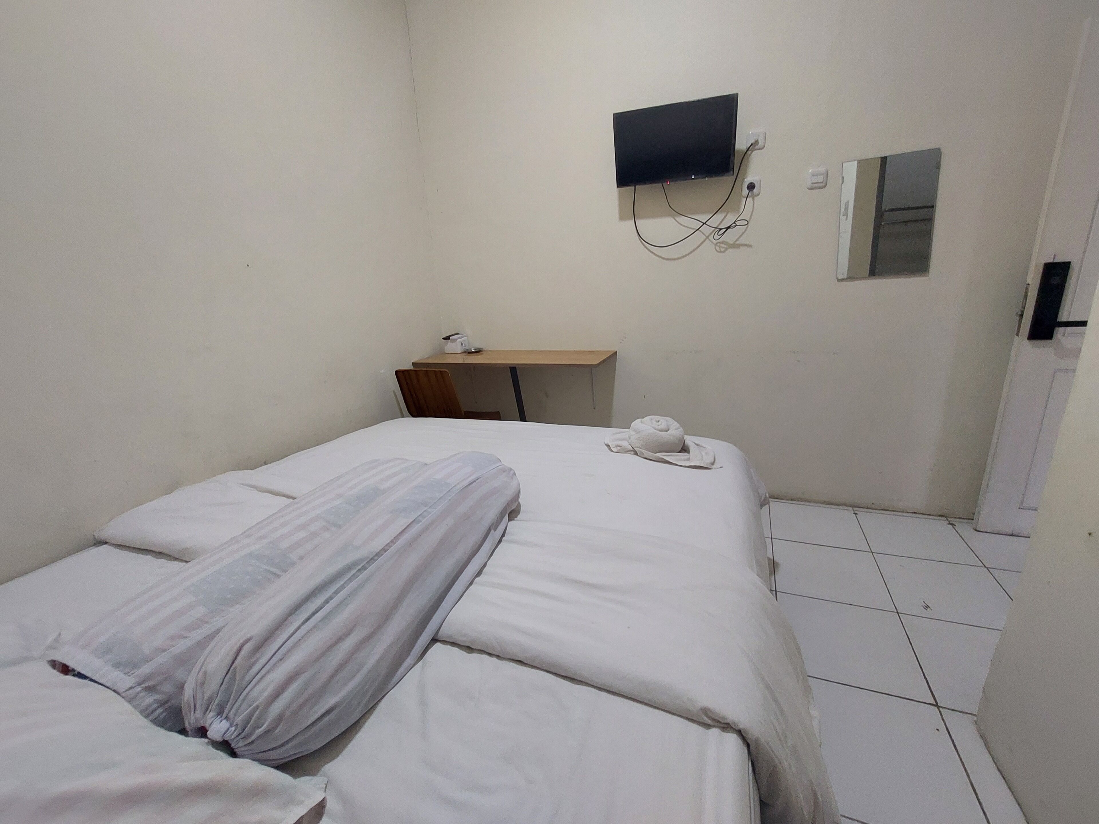 A small, sparsely furnished room with a bed, a desk, and a TV mounted on the wall.