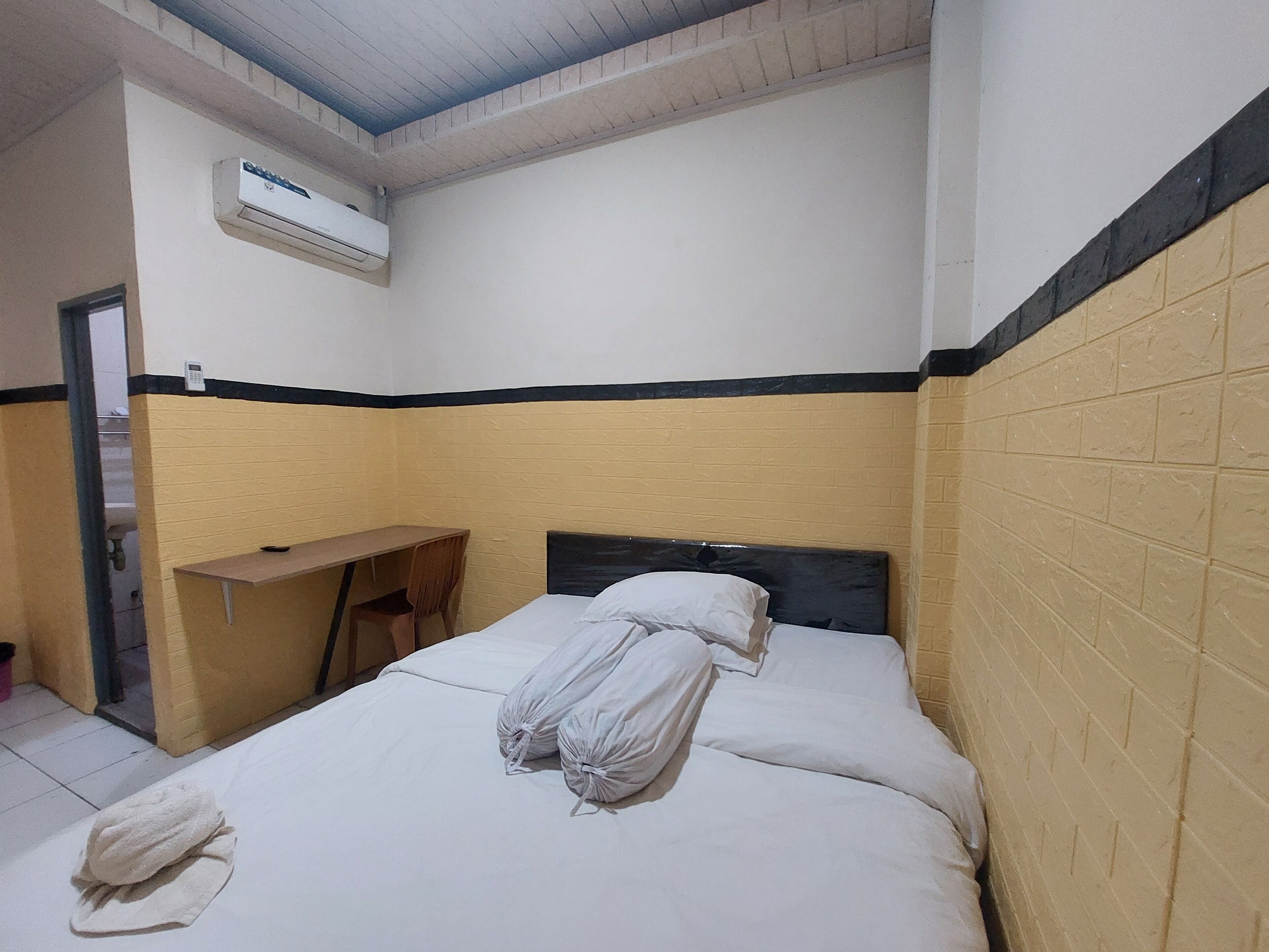 A single bed with a white mattress and pillows, a wooden desk, and an air conditioning unit mounted on the wall.